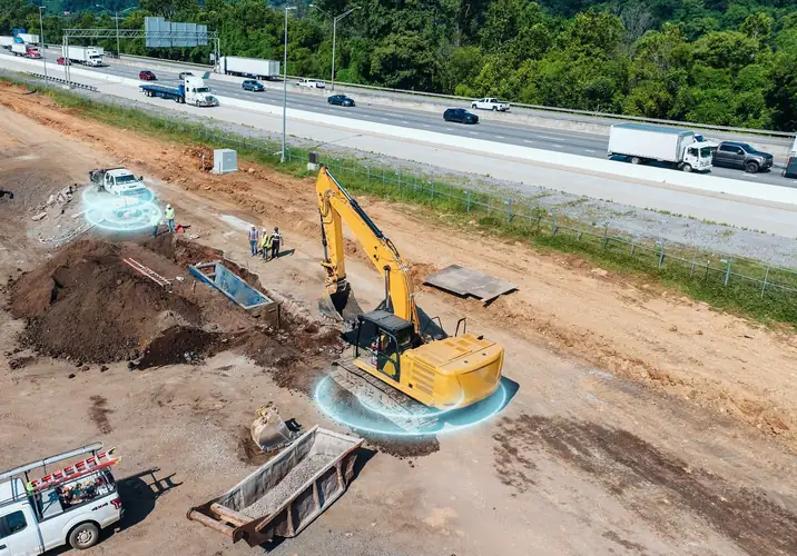 A trench job beside a highway: an excavator digging, a crew in hi-vis, a plate compactor and a service ute